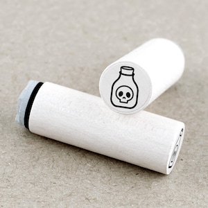Op de afbeelding: Twee witte houten stempels. Eén stempel toont een zwarte omtrek van een fles met een schedel erin. De andere stempel staat op de achtergrond. De stempels zijn cilindrisch met een grijs stempelkussen aan één uiteinde.
