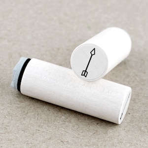 Op de afbeelding: Twee witte houten stempels met een zwart pijldesign. Eén stempel ligt bovenop de andere. De pijl is een eenvoudige lijntekening met een puntige punt en een bevederde basis.
