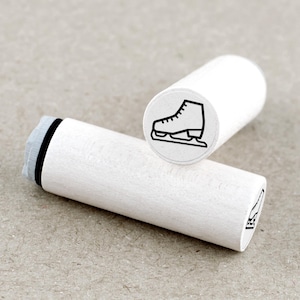 Op de afbeelding: Twee witte houten stempels met een zwarte illustratie van een schaats. Eén stempel staat rechtop en toont de schaatsafbeelding, terwijl de andere horizontaal ligt. De stempels zijn cilindrisch en hebben een eenvoudig, strak ontwerp.