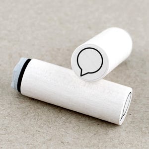 Könnte beinhalten: Zwei weiße Holzstempel mit einem Sprechblasen-Design. Die Stempel haben eine zylindrische Form mit einem schwarzen Sprechblasen-Umriss auf der Oberseite. Ein Stempel liegt auf dem anderen.