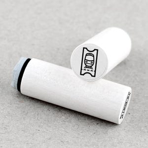 May include: Two white wooden stamps. One stamp features a black and white train ticket design. The other stamp has a black border and a grey stamp pad. The stamps are cylindrical and have a minimalist design.