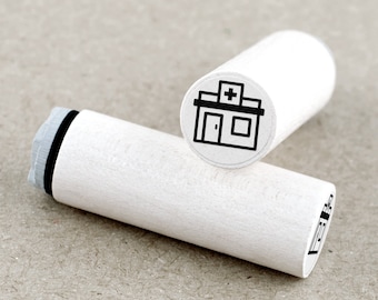 May include: Two white wooden stamps with a black illustration of a building with a cross on top, likely a hospital or clinic. The stamps are cylindrical with a black ink pad visible on one end.