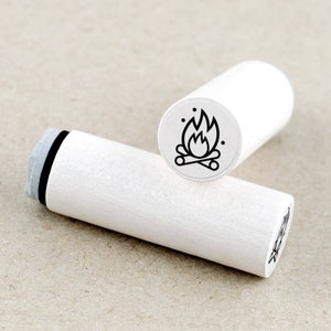 Könnte beinhalten: Weißer Holzstempel mit einer schwarzen Linienzeichnung eines Lagerfeuers mit Flammen. Der Stempel ist zylindrisch und hat eine schwarz-weiße Oberseite.