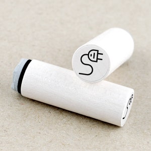 May include: White wooden stamp with a black outline of an electrical plug and cord. The stamp is round and has a raised edge.
