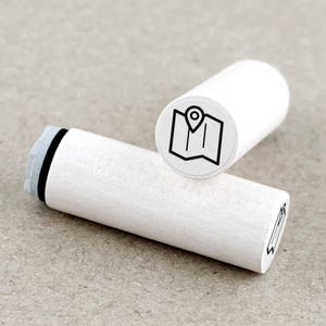 Op de afbeelding: Twee witte houten stempels met een kaart- en locatie-pin-icoon. Eén stempel staat rechtop en toont het icoon, terwijl de andere horizontaal ligt. De stempels zijn cilindrisch met een grijs inktkussen aan één uiteinde. De achtergrond is een neutraal, lichtbruin.