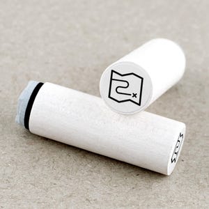 Op de afbeelding: Twee witte houten stempels met zwarte inktontwerpen. Eén stempel toont een kaart met een route en een X die een locatie markeert. De andere stempel heeft een vergelijkbaar ontwerp. De stempels zijn cilindrisch en hebben een lichtgrijs inktkussen.