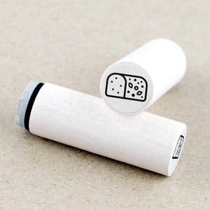 Könnte beinhalten: Zwei weiße Holzstempel mit einem schwarzen Brotlaib-Design. Ein Stempel steht aufrecht und zeigt die Brotillustration, während der andere horizontal liegt. Das Brot-Design umfasst eine Kruste, Samen und ein gepunktetes Muster.