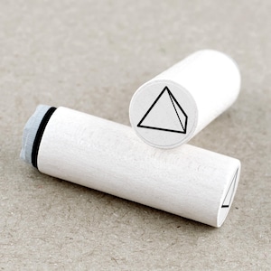 May include: Two white wooden stamps with a geometric pyramid design. The stamps have a cylindrical shape with a black pyramid outline on the circular stamp face. One stamp is positioned on top of the other.