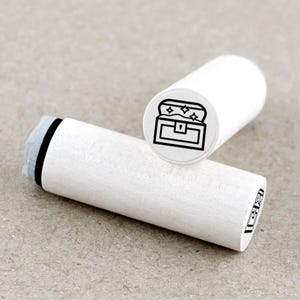 Op de afbeelding: Twee witte houten stempels met een zwarte omtrekillustratie van een open schatkist met sterren. Eén stempel staat rechtop en toont het kistontwerp, de andere ligt horizontaal.