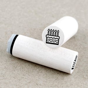 Peut inclure: Deux tampons en bois blancs avec un motif de gâteau d'anniversaire noir. L'un des tampons est droit, affichant le gâteau avec cinq bougies. L'autre tampon est couché, montrant le manche en bois et le motif du tampon.