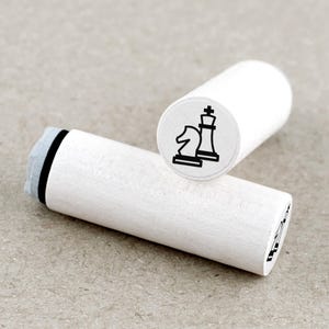 Op de afbeelding: Twee witte houten schaakstempels. Eén stempel toont een zwart paard- en koningschaakstukontwerp. De andere stempel toont hetzelfde ontwerp. De stempels zijn cilindrisch met een grijs inktkussen aan één uiteinde.