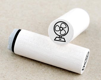 May include: Two white wooden stamps with a black outline illustration of a fan. One stamp is upright, displaying the fan image, while the other lies horizontally. The stamps are cylindrical and have a simple, clean design.