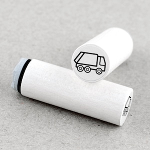May include: Two white wooden stamps with a black outline of a garbage truck. One stamp is upright, displaying the truck image, while the other lies horizontally. The stamps are cylindrical with a gray and black ink pad.