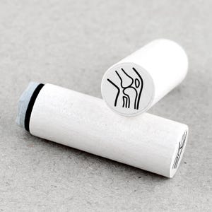 Op de afbeelding: Twee witte houten stempels met een zwarte illustratie van een kniegewricht. Eén stempel staat rechtop en toont de afbeelding van het kniegewricht, terwijl de andere horizontaal ligt. De stempels zijn cilindrisch.