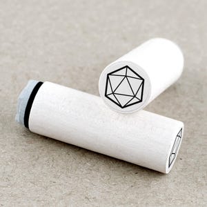 Könnte beinhalten: Zwei weiße Holzstempel mit einem geometrischen Design. Ein Stempel zeigt das Design am Ende, eine schwarze Umrisslinie einer vielseitigen Form. Der andere Stempel liegt, zeigt das geometrische Design am Ende und einen Teil des Stempeldesigns.