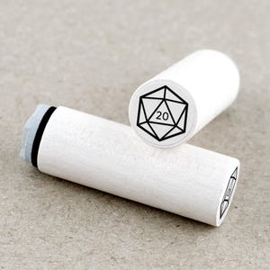 May include: Two white wooden stamps. One stamp shows a black and white twenty-sided die with the number 20. The other stamp has a similar design. The stamps are cylindrical with a gray ink pad.