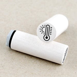 Könnte beinhalten: Zwei weiße Holzstempel mit einem schwarzen Thermometer- und Sonnendesign. Ein Stempel steht aufrecht und zeigt das Design, während der andere horizontal liegt. Die Stempel sind zylindrisch mit einem grauen Stempelkissen an einem Ende. Das Design deutet auf ein Wetter- oder Temperaturthema hin.