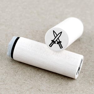 Op de afbeelding: Twee witte houten stempels met een zwart ontwerp van gekruiste zwaarden. De stempels zijn cilindrisch met een plat, cirkelvormig stempelvlak. Eén stempel staat rechtop en toont het ontwerp, de andere ligt horizontaal.