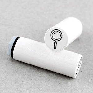 Peut inclure: Deux tampons en bois blancs avec un motif de loupe noire. L'un des tampons est droit, affichant la loupe, tandis que l'autre est horizontal. Les tampons sont cylindriques et présentent une esthétique simple et épurée.