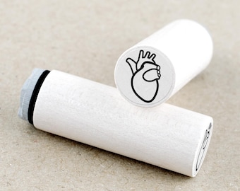 May include: Two white wooden stamps with a black heart illustration. One stamp is upright, displaying the heart design, while the other lies horizontally. The stamps are cylindrical with a gray and black ink pad.