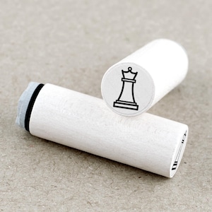 Op de afbeelding: Twee witte houten schaakstempels. Eén stempel toont een zwart schaakstuk van een koningin. De andere stempel toont een zwart schaakstuk van een koningin. De stempels zijn cilindrisch en liggen op een neutraal gekleurd oppervlak.