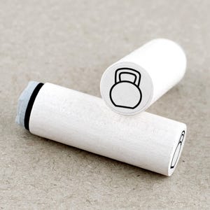 Op de afbeelding: Twee witte houten stempels met zwarte inktkussens. Een stempel heeft een lijntekening van een kettlebell.