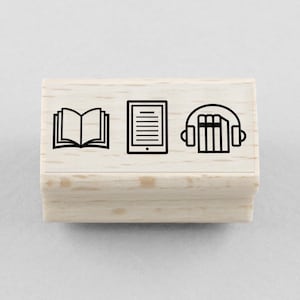 Può includere: Un timbro in legno con tre icone nere: un libro aperto, un tablet e cuffie con libri. Il timbro è rettangolare e realizzato in legno chiaro. Le icone rappresentano la lettura, gli e-book e gli audiolibri.