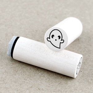 Könnte beinhalten: Zwei weiße Holz-Stempel mit schwarzen Stempelkissen. Der Stempel links ist leer. Der Stempel rechts hat eine Schwarzweiß-Illustration eines Geistes mit Augen und Mund.