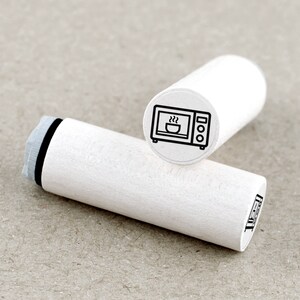 Könnte beinhalten: Weißer Holzstempel mit einer schwarzen Linienzeichnung einer Mikrowelle, aus der Dampf aufsteigt. Der Stempel ist etwa 5 cm lang.