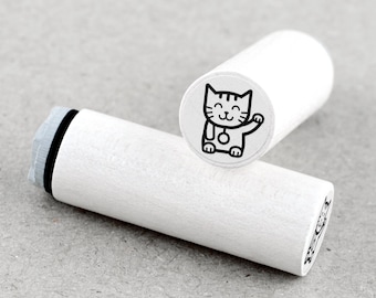 Op de afbeelding: Witte houten stempel met een zwarte lijntekening van een kat met een bel om zijn nek. De kat glimlacht en zwaait met zijn poot.