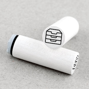 Op de afbeelding: Twee witte houten stempels met zwarte inktontwerpen. Eén stempel toont een kast met drie laden, de andere drie gestapelde rechthoeken. De stempels zijn cilindrisch en hebben een grijs inktkussen.