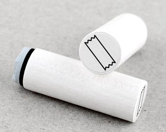 May include: Two white wooden stamps with a black tape design. One stamp is upright, and the other is lying down. The stamps have a cylindrical shape and are set against a gray background. The tape design is a simple, diagonal rectangle with a zig-zag edge.