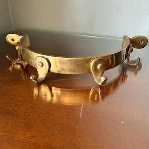 May include: A curved brass wall-mounted rack featuring four individual hooks attached to a central semi-circular bar. Each hook is secured with a small copper-toned rivet, and the ends of the bar feature flat mounting plates with screw holes.