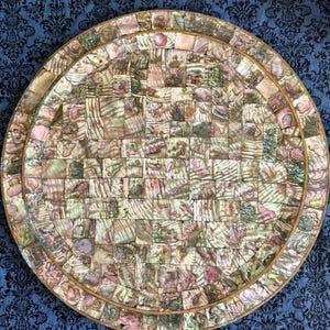 May include: A circular decorative tray featuring a mosaic pattern composed of small, square iridescent abalone shell tiles. The surface displays a variety of shimmering pink, green, and cream tones, framed by a thin, light-colored wooden border.