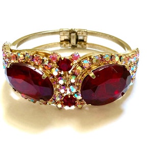 May include: A gold tone hinged bangle bracelet with two large red oval-shaped stones surrounded by smaller clear and pink rhinestones.