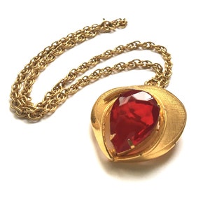 May include: A gold chain necklace with a large red teardrop-shaped rhinestone pendant. The pendant is set in a gold frame with a curved design.