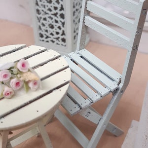 May include: A light blue wooden folding chair and a white wooden table with a bouquet of pink flowers on top. The table and chair are both distressed and have a rustic look.