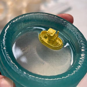 Pode incluir: Um pequeno barco de brinquedo amarelo flutua numa substância clara, semelhante à água, dentro de um recipiente circular turquesa. O recipiente tem uma borda texturizada e ondulada. O barco tem uma cabine e outros detalhes.