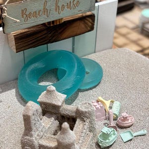 以下が含まれることがあります： 砂のお城、ターコイズ色の浮き輪、パステルカラーの小さなおもちゃが置かれたミニチュアビーチのシーン。 「Beach house」と書かれた木製の看板が上に吊り下げられており、遊び心のある夏のテーマを連想させます。