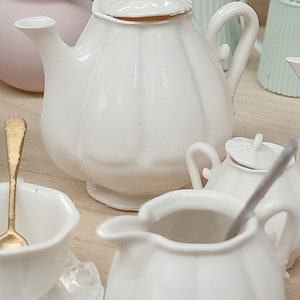 Könnte beinhalten: Eine weiße Porzellan-Teekanne mit Deckel, ein weißer Milchkännchen und eine weiße Zuckerdose mit Deckel. Die Teekanne hat einen runden Körper und einen gebogenen Ausguss. Der Milchkännchen und die Zuckerdose haben einen runden Körper und einen kleinen Griff.