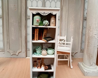 May include: A white bookshelf with five shelves, filled with various decorative items, including books, jars, cups, candlesticks, and a hat. The bookshelf is in a room with a white chair and a white pillar.