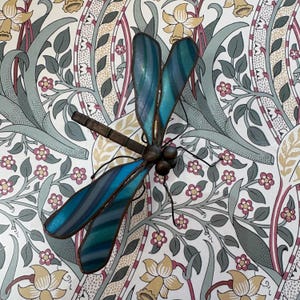May include: A decorative dragonfly sculpture with blue and green iridescent wings. The dragonfly has a dark brown body and wire legs. The artwork is mounted on a floral wallpaper background with a repeating pattern.