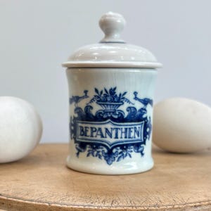 May include: A white ceramic apothecary jar with a lid, decorated with blue floral and bird motifs. The jar displays the word "BEPANTHEN" in a blue banner. The jar is on a wooden surface.