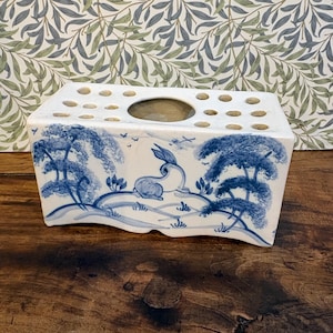 May include: A white ceramic flower frog with a blue and white landscape design. The design features a rabbit, trees, and birds. The top has multiple holes for arranging flowers. The frog is on a wooden surface.