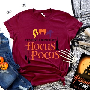 May include: A maroon short-sleeved t-shirt featuring the text "IT'S JUST A BUNCH OF HOCUS POCUS" in orange and black, accompanied by silhouettes of three hairstyles and two broomsticks, paired with blue denim jeans and a Halloween party card.