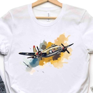 May include: White t-shirt with a watercolor illustration of a vintage military aircraft. The plane is painted in camouflage colors and has a large propeller. The illustration is surrounded by a splash of yellow and orange watercolor.