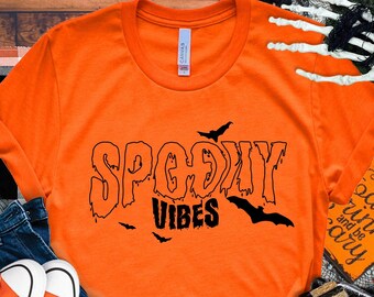 Op de afbeelding: Een oranje T-shirt met korte mouwen en de zwarte tekst SPOOKY VIBES in een druipend lettertype, vergezeld van zwarte vleermuissilhouetten. Het T-shirt wordt getoond naast een blauwe spijkerbroek en oranje canvas sneakers.