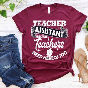 K&ouml;nnte beinhalten: Ein maroonfarbenes T-Shirt mit dem Text "Teacher Assistant Because Teachers Need Heros Too" in Wei&szlig; mit einer schwarzen Apfelgrafik.