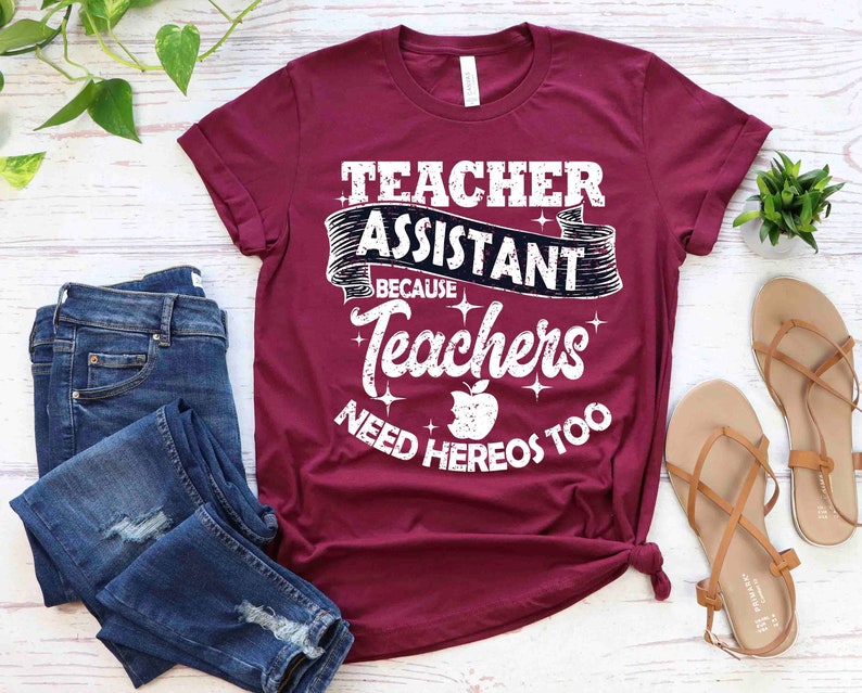 K&ouml;nnte beinhalten: Ein maroonfarbenes T-Shirt mit dem Text "Teacher Assistant Because Teachers Need Heros Too" in Wei&szlig; mit einer schwarzen Apfelgrafik.