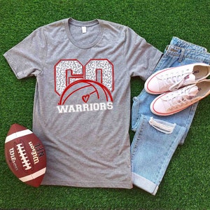 Puede incluir: Camiseta gris con un gr&aacute;fico rojo y blanco de un bal&oacute;n de voleibol con el n&uacute;mero "60" en un dise&ntilde;o de estampado de leopardo y el texto "WARRIORS".